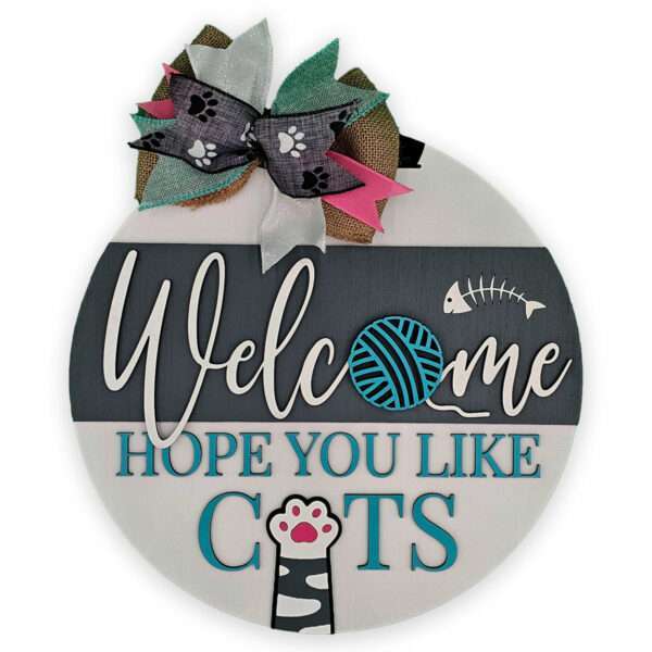 A round wooden sign with three sections. The top has a decorative bow with paw patterns. The middle section reads "Welcome" with a yarn ball and fishbone design. The bottom section says "Hope you like cats" with a cat paw and a turquoise background.