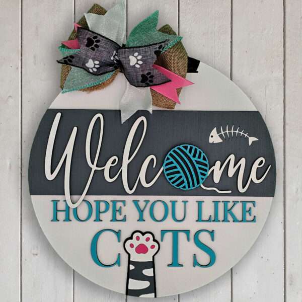 Round wooden welcome sign against a white wooden background. The sign reads "Welcome, Hope You Like Cats" with a yarn ball and fish skeleton replacing the "o" in "Welcome." A bow with paw print ribbons is at the top and a cat paw print at the bottom.