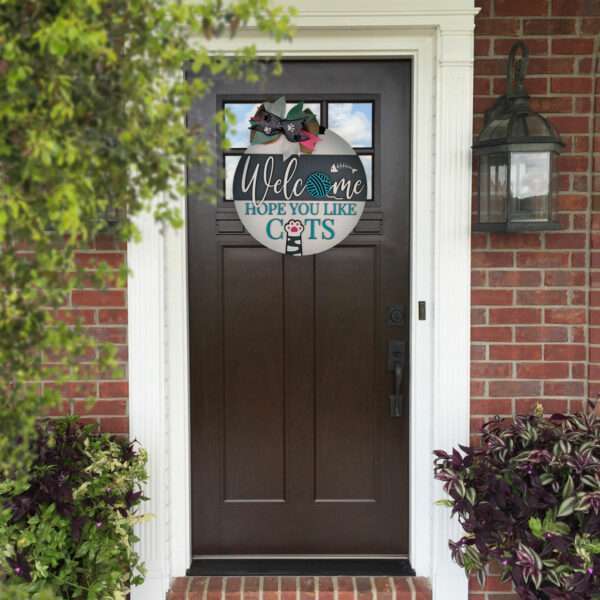 A dark brown front door with a custom door sign that reads "Welcome, Hope You Like Cats" hanging in the center. The door is framed by a brick exterior and two large plants on either side. A vintage-style lantern is mounted on the brick to the right of the door.