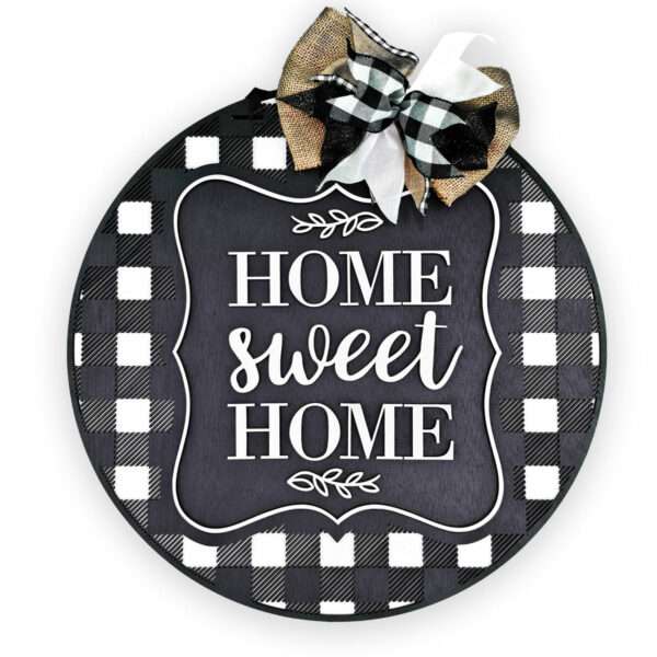 A round sign with a black and white buffalo plaid background features the phrase "Home Sweet Home" in white lettering. Adorning the sign is a decorative black, white, and gold plaid ribbon bow at the top.