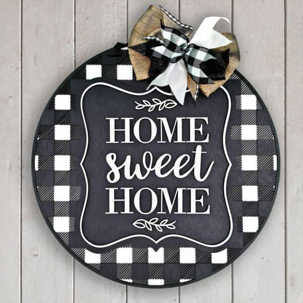 A round sign with a black and white buffalo check pattern border and a chalkboard-style center reads "Home Sweet Home" in white scripted text. The sign is adorned with black, white, and gold ribbons tied at the top. The sign hangs on a wooden wall.