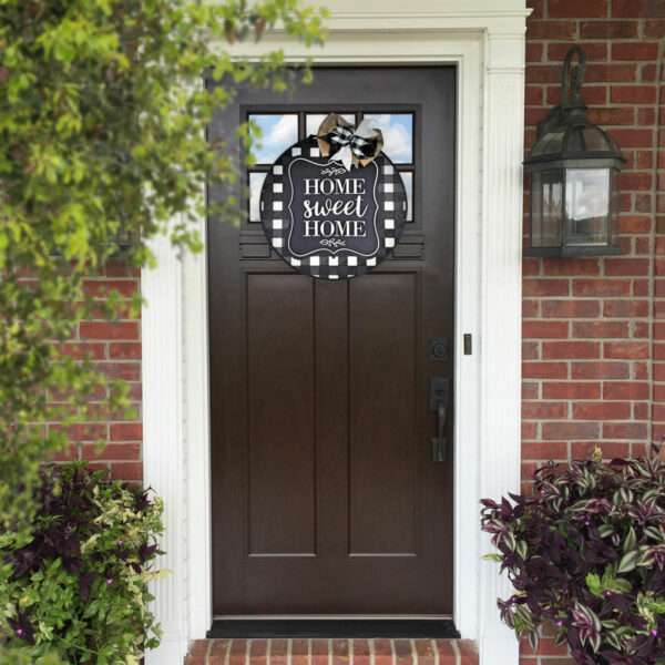 A dark brown front door with a custom "Home Sweet Home" sign hanging on it. The door is set in a red brick exterior with white trim and has a black outdoor lantern on the right. Flanking the door are green plants and shrubs.