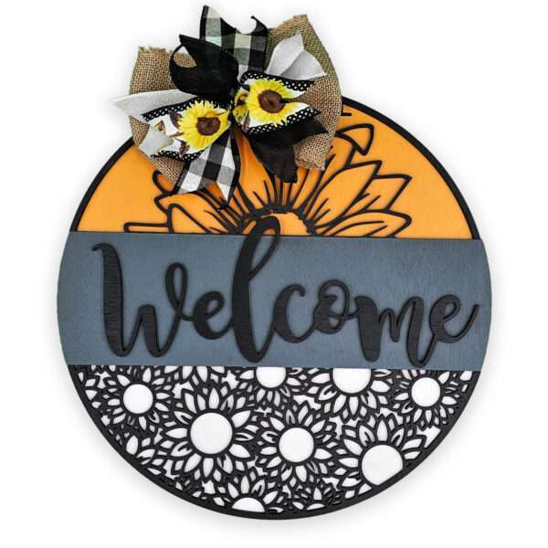 A round, decorative welcome sign featuring a large "welcome" text across the center. The top section has a large orange sunflower design, the bottom section has a black and white sunflower pattern. The sign is adorned with a black and white checkered bow with sunflowers.