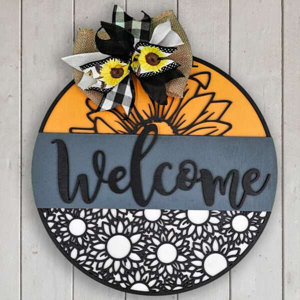 A round wooden welcome sign designed with an orange top section, a dark gray middle band with the word "welcome" in black cursive letters, and a white bottom section with a sunflower pattern. A bow with sunflower print and black-and-white gingham ribbon is at the top.