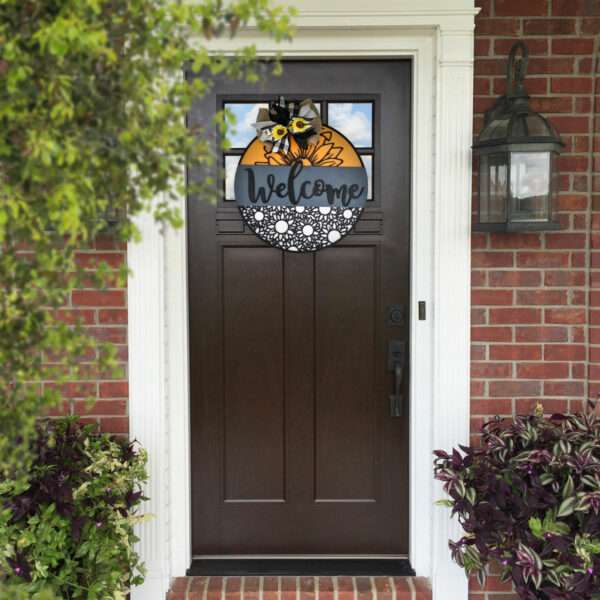 A wooden front door with a custom "Welcome" sign featuring a sunflower and a bee is centered in the image. The door has a small rectangular window at the top, and there is a lantern-style light fixture on the right side. Potted plants are placed on both sides of the entrance.