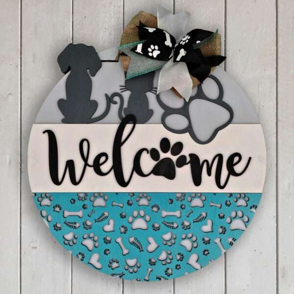 Introducing the "Welcome Cats and Dogs" sign by Rustic Road Creations. This round wooden sign, featuring a "Welcome" message in cursive, is adorned with silhouettes of a dog and a cat, paw prints, bones, and hearts. The top is highlighted by a bow with paw print designs. The background showcases light blue and aqua tones with paw print patterns.