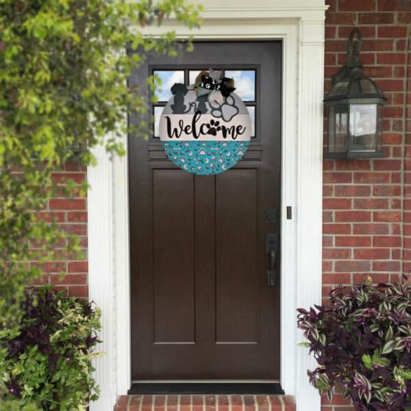 A brown front door with a white frame is decorated with a round "Welcome Cats and Dogs" sign from Rustic Road Creations, featuring cat and dog silhouettes in blue. The door is surrounded by red brick walls, a black wall lantern on the right, and potted plants with purple and green leaves on both sides.