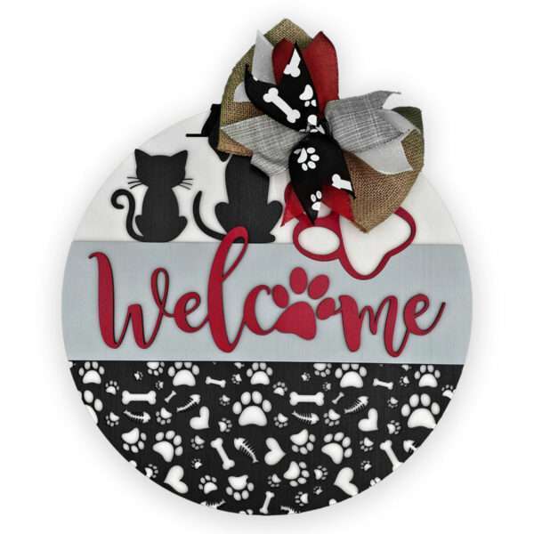 Introducing the "Welcome Cats and Dogs" sign by Rustic Road Creations. This circular welcome sign features a black cat silhouette, a dog silhouette, and a red paw print on a white background. The word "Welcome" is written in striking red with a paw print replacing the letter "o." The bottom section showcases an engaging pattern of bones and paw prints, topped off with an elegant large bow.