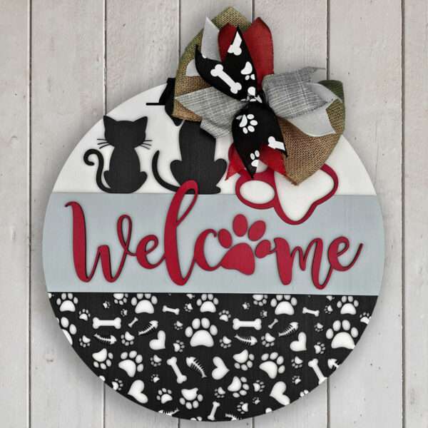 A round wooden sign from Rustic Road Creations' "Welcome Cats and Dogs" collection hangs on a white wall, showcasing the silhouettes of a cat and a dog adorned with a large bow atop. The word "Welcome" is prominently displayed in red script, featuring a paw print as the "o." The bottom section is decorated with an intricate pattern of bones, paws, and hearts.