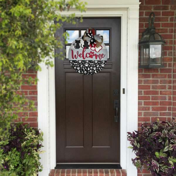 A red front door adorned with the "Welcome Cats and Dogs" wreath by Rustic Road Creations, featuring floral and ribbon decorations. The door is part of a red brick house, flanked by a lantern on the right and potted plants on both sides.