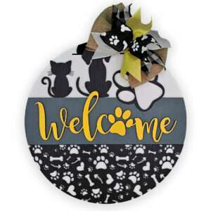 The "Welcome Cats and Dogs" round door sign by Rustic Road Creations is a yellow sign featuring the word "Welcome" with a paw print replacing the letter "o." The top half displays silhouettes of two cats and a large paw print, while the bottom half showcases a pattern of various pet-related icons. A ribbon bow is attached at the top for added charm.