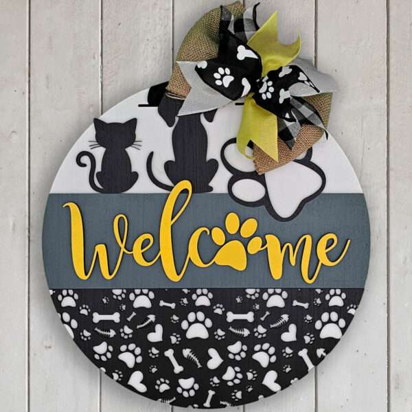 Round decorative sign by Rustic Road Creations, named "Welcome Cats and Dogs," adorned with a bow at the top left corner and featuring silhouettes of cats, dog paws, and dog bones. The word "Welcome" is prominently displayed in yellow with a paw print incorporated within the text, against a background of paw and bone patterns.