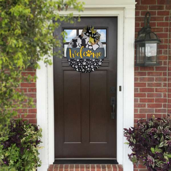 A brown front door adorned with a circular welcome sign featuring the "Welcome Cats and Dogs" design by Rustic Road Creations in yellow. The door is framed by white trim and red brick walls. On either side of the entrance are potted plants with dark purple and green foliage. A lantern-style light fixture is mounted to the right.