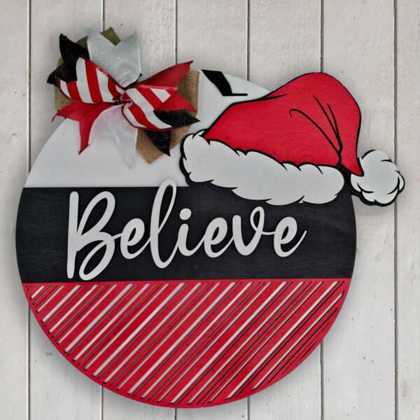 A round Christmas sign hangs on a wooden wall. The sign's top half features the words "Believe in Santa" in white, written on a black background, with a Santa hat graphic on the upper right corner. The bottom half has diagonal red and white stripes. A festive bow is at the top left.