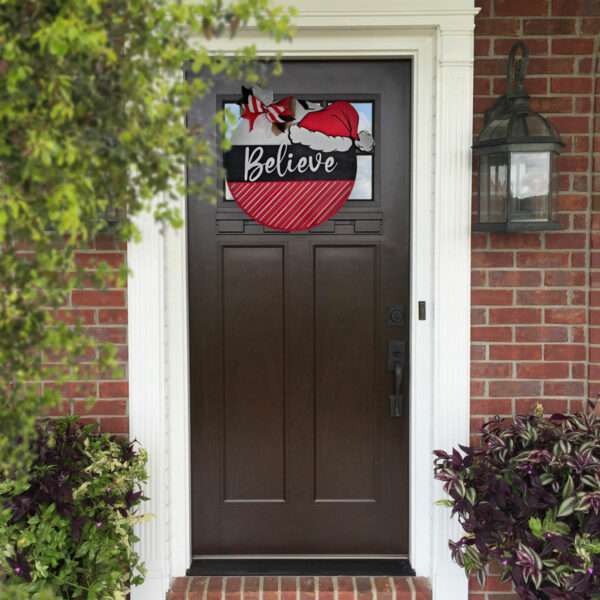 A front door adorned with a custom Christmas sign. Shaped like a round ornament, it features the words "Believe in Santa" along with images of his hat and belt. The door, framed by white trim and red brick walls, is complemented by plants on either side.