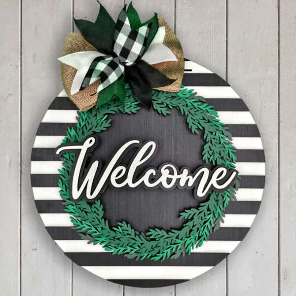 A decorative round door sign with black and white horizontal stripes. It features a green wreath and the word "Welcome" in the center. At the top is a bow made of black and white buffalo plaid and burlap ribbons. The background is a light wooden panel.