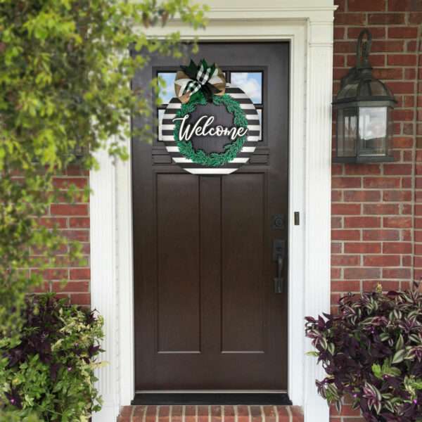 A dark brown front door with a circular green and black "Welcome" wreath is centered in a brick house entrance. A black, lantern-style light fixture is mounted beside the door, next to a custom door sign. Surrounding the entrance are green shrubs and purple-leaved plants.