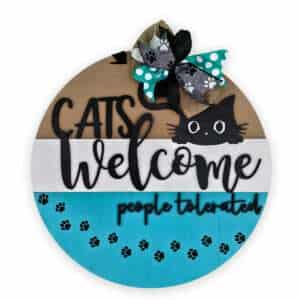 A round wooden sign with decorative elements reads "Cats Welcome, People Tolerated." The sign features a black cat's head peeking over the middle section, a paw print pattern on the lower section, and a multi-patterned ribbon bow at the top.