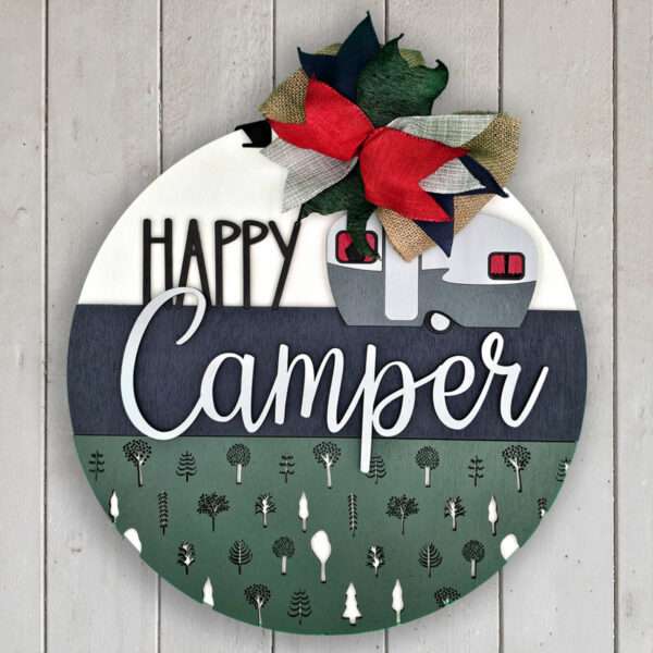 A decorative round sign features the text "Happy Camper" with a camper illustration and pine trees below. A bow made of red, green, and burlap fabric adorns the top left of the sign. The background is made of wooden slats.