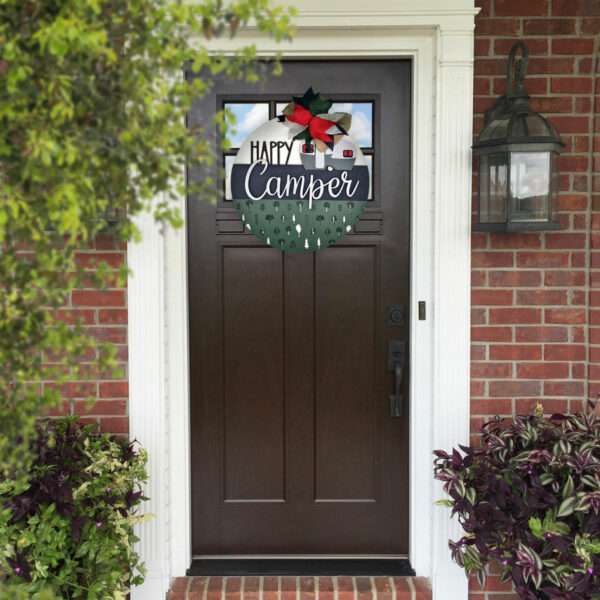 A dark brown front door with a custom sign hanging on it that reads "Happy Camper" in a mix of white and green letters, adorned with a bow and holly leaves. The door is set in a white frame with a brick wall backdrop. Plants are on either side of the door.