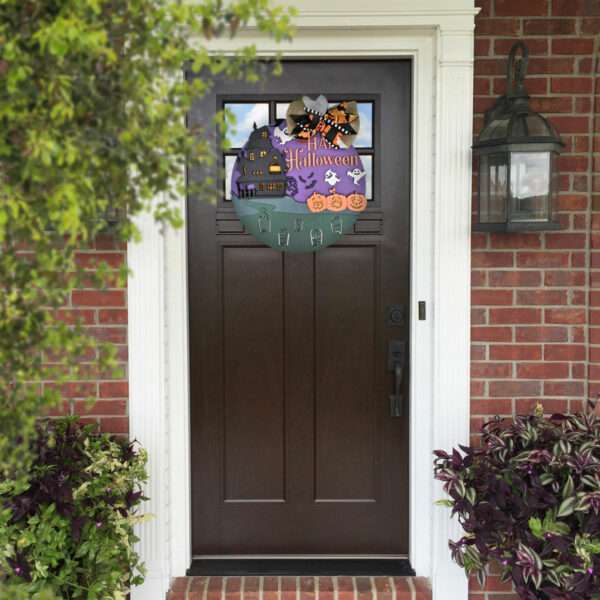 A brown front door adorned with a colorful Halloween wreath featuring haunted houses, bats, ghosts, and pumpkins. The door is set in a brick wall, flanked by a large lantern-style light fixture on the right, potted plants on both sides, and a custom "Happy Halloween" door sign adding festive charm.