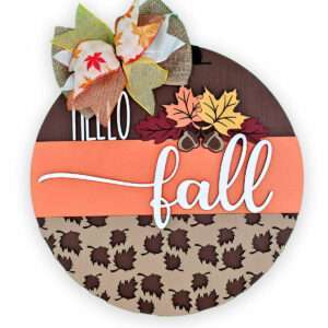 A custom door sign reads "Hello fall" in white letters on a round wooden base. The background features three sections: dark wood, orange, and beige with leaf patterns. Topped with a decorative bow of autumn leaves, the design is further enhanced by two acorns and additional autumn leaves.