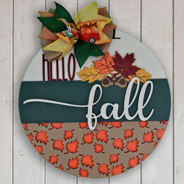 A round, fall-themed custom door sign features "Hello Fall" written on it. The top third is green with a plaid bow and pumpkin detail. The middle has colorful fall leaves. The bottom section is patterned with red and orange leaves on a beige background.