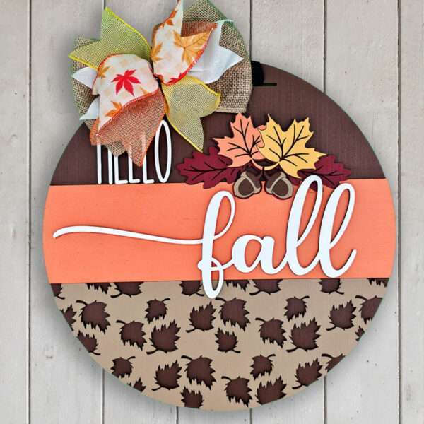 A decorative round wooden custom door sign with a fall theme. The top section says “HELLO” in white letters, and the middle section says “fall” in white cursive. It is adorned with autumn leaves, acorns, and a colorful bow with leaf patterns on the upper left. Perfect for saying hello fall!