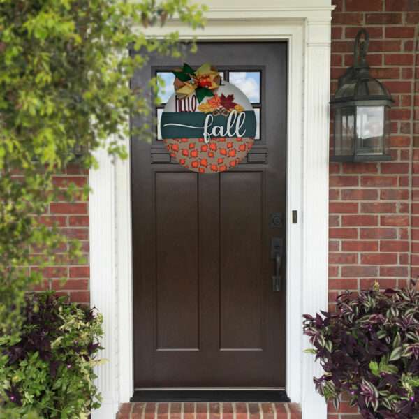 A dark brown front door with a custom door sign that says "Hello Fall" hangs in the center. The fall-themed wreath is decorated with leaves and a bow. The entrance is framed by red bricks and two white columns, while plants and a lantern are visible nearby.