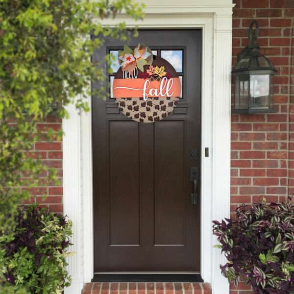 A dark brown front door is adorned with a circular autumn wreath featuring leaves, flowers, and an orange ribbon that reads "hello fall." The entrance, framed by white columns, is surrounded by green and purple plants. A custom door sign hangs beside the brick wall and lantern to the right.