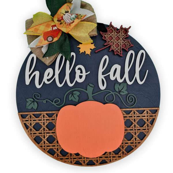 A circular custom door sign features the text "hello fall" in white cursive letters. It is decorated with an orange pumpkin, a yellow bow adorned with a small truck and pumpkin, and a red maple leaf cutout. The bottom part includes a pattern resembling a wooden lattice.