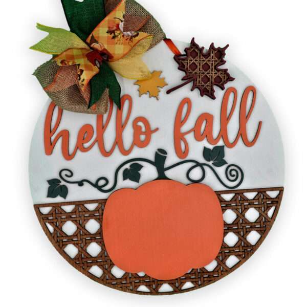 A custom door sign featuring the phrase "hello fall" in orange cursive letters, adorned with a pumpkin, leaf cutouts, and a patterned bottom half. A burlap bow with autumnal colors is attached to the top left corner.