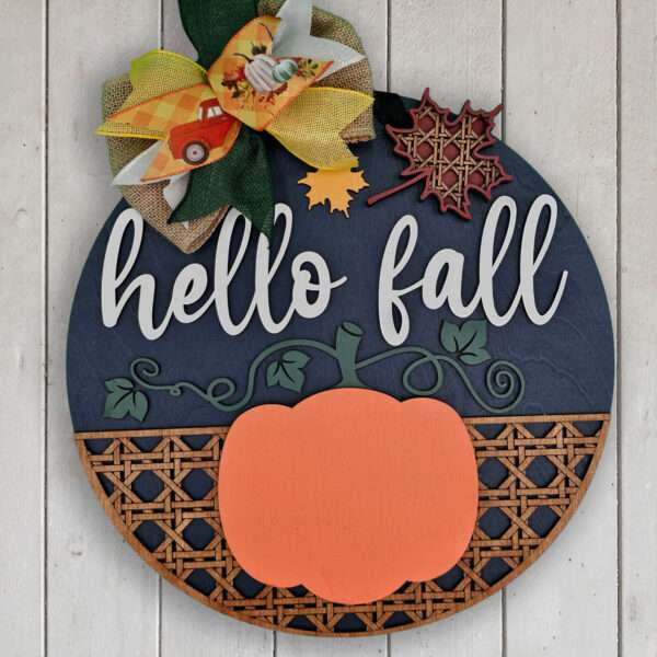 A round wooden custom door sign with "hello fall" written in orange cursive. It is decorated with an orange pumpkin and autumn leaves, including a 3D-style maple leaf. A burlap bow accented with plaid and solid autumn colors is attached at the top left, perfect for welcoming the season.