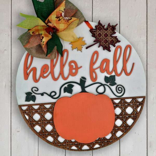 A custom door sign features a round wood design against a white background with "hello fall" written in orange cursive. Below the text is a delightful hello fall pumpkin graphic. The top boasts a large, colorful bow and small autumn leaves, while the bottom showcases an elegant lattice pattern.