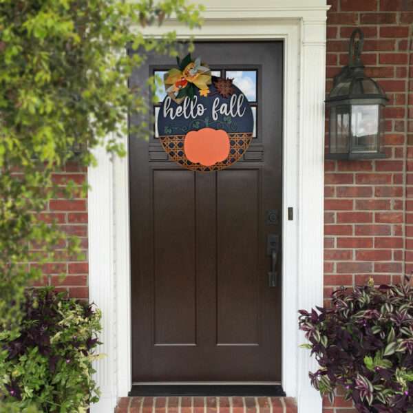 A dark door with a custom door sign reading "Hello Fall" hangs on it, adorned with a fall-themed wreath featuring leaves and a pumpkin. The door is framed by white trim, brick siding, and flanked by potted plants and outdoor lanterns. Green foliage partially frames the image on the left.