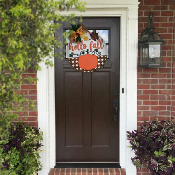 A dark brown door with white trim features a fall-themed wreath adorned with leaves, pine cones, and a hello fall pumpkin sign. The brick wall around the door has a lantern light fixture on the right and greenery on both sides.