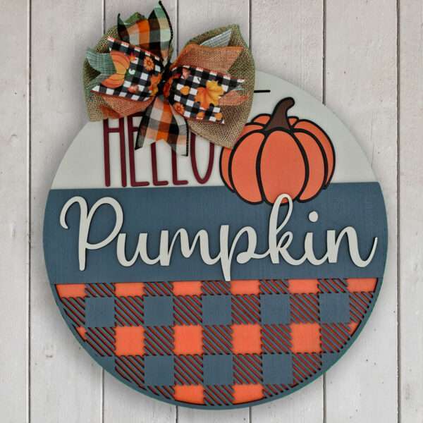 A round wooden sign with "HELLO Pumpkin" written on it. The top part features "HELLO" in red and an orange pumpkin graphic. Below, "Pumpkin" is written in white cursive on a blue and orange checkered background while a plaid bow decorates the top, making it a warm welcome for fall.