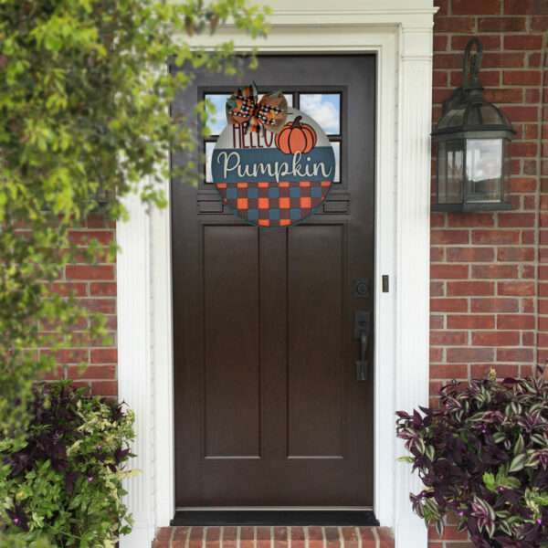 A dark brown front door of a brick house features a charming custom door sign that reads "Hello Pumpkin," adorned with pumpkins and a checkered pattern. White columns, a black lantern light fixture on the right, and potted plants with green and purple leaves flank the welcoming entrance.