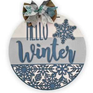 A custom door sign, round and wooden, is adorned with a bow made of gray, white, and teal fabric. Divided into three sections with varying shades of blue and decorated with snowflakes, the centerpiece prominently displays the text "Hello Winter.