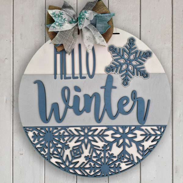 A custom door sign, round and wooden, is adorned with a bow made of gray, white, and teal fabric. Divided into three sections with varying shades of blue and decorated with snowflakes, the centerpiece prominently displays the text "Hello Winter. The sign is placed on a white wood backgorund.