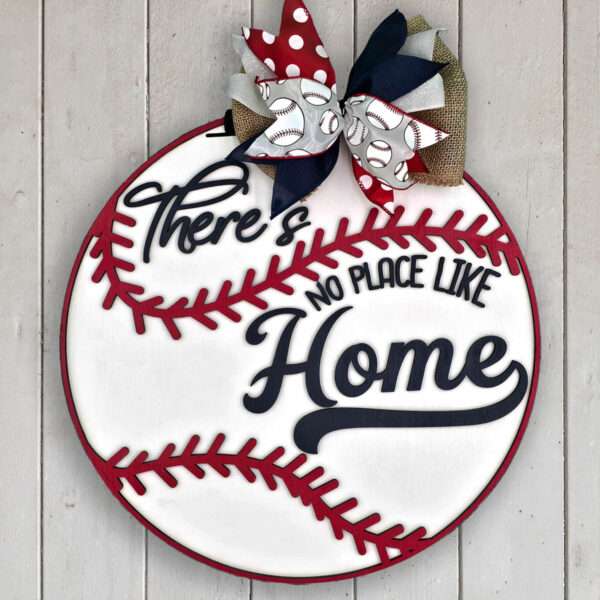 A round baseball-themed sign reads "There's no place like home" in stylized text. The sign is adorned with a decorative bow featuring red, white, and blue ribbons, some with baseball patterns. This charming piece hangs elegantly on a light wooden wall.