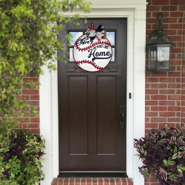 A brown front door on a brick house is adorned with a custom door sign resembling a baseball, featuring the text "There's no place like home" in black and red lettering. Green bushes and plants frame the doorway, while a wall-mounted lantern adds charm to the right of the entrance.