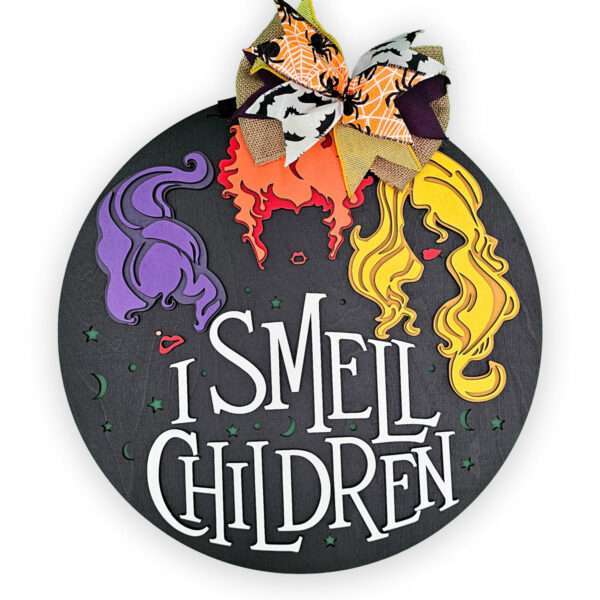 A round black sign with the bold white text "I Smell Children." Above it, you'll find stylized illustrations of three hair silhouettes: one purple, one orange, and one yellow. The sign is beautifully adorned with a bow at the top, showcasing a vibrant mix of autumn-themed colors and patterns.