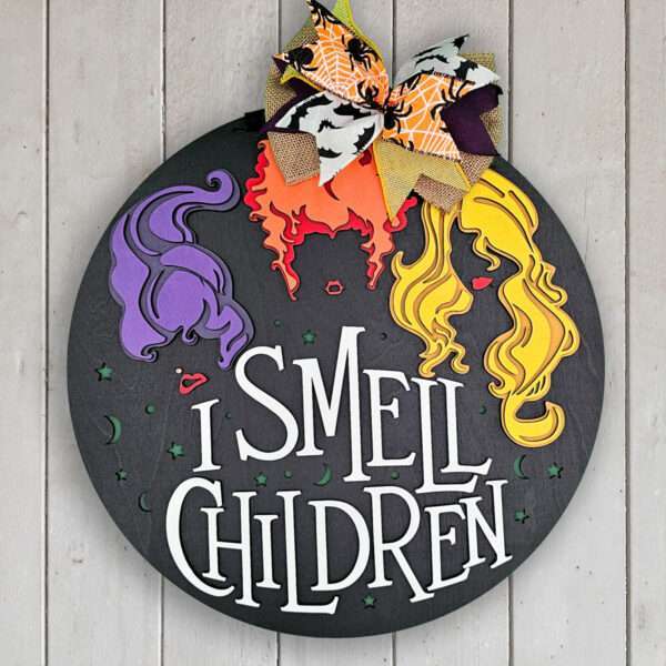 A round black wooden sign, inspired by Hocus Pocus, features three colorful hairstyles - one purple, one orange, and one yellow - above the text "I SMELL CHILDREN" in white. A decorative bow with orange, black, white, and plaid patterns is attached at the top. The background is light wood planks.