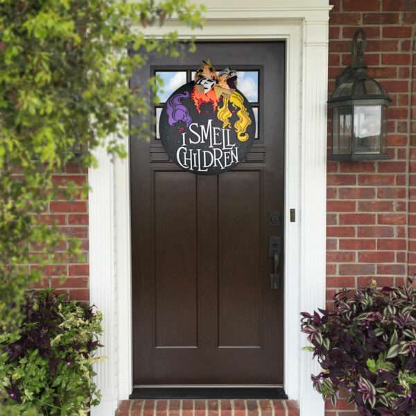 A front door with a dark wood finish features a custom door sign, displaying three silhouetted witches' heads and the text, "I Smell Children." The door is framed by white columns and brick walls, with leafy plants on either side.