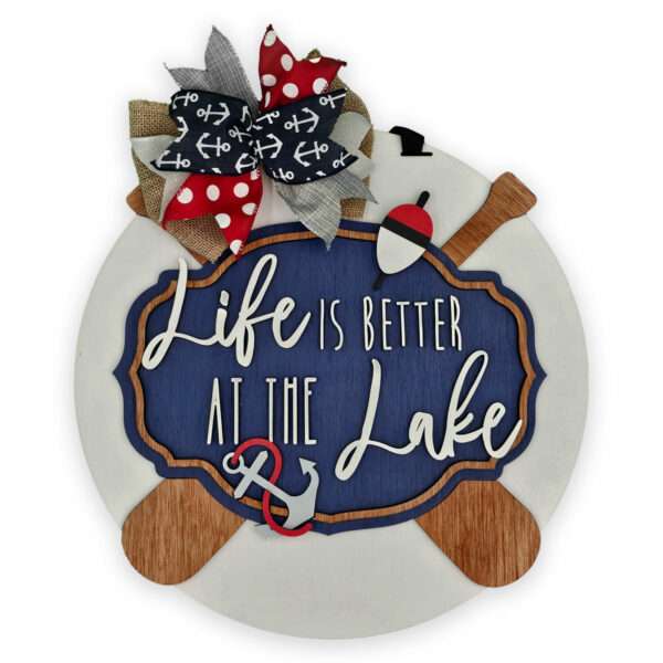 A round decorative sign with wooden paddles at the bottom and a colorful bow at the top, featuring a life preserver and anchor designs. The center has a navy blue background with white and cursive text that reads, "Life is better at the Lake.