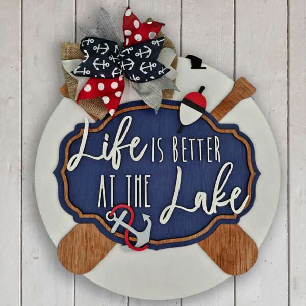 A round wooden sign with a navy blue center reads "Life is better at the lake" in white script. It is decorated with a large burlap and ribbon bow, wooden oars, and an anchor. The sign is mounted on a white, rustic wooden background.