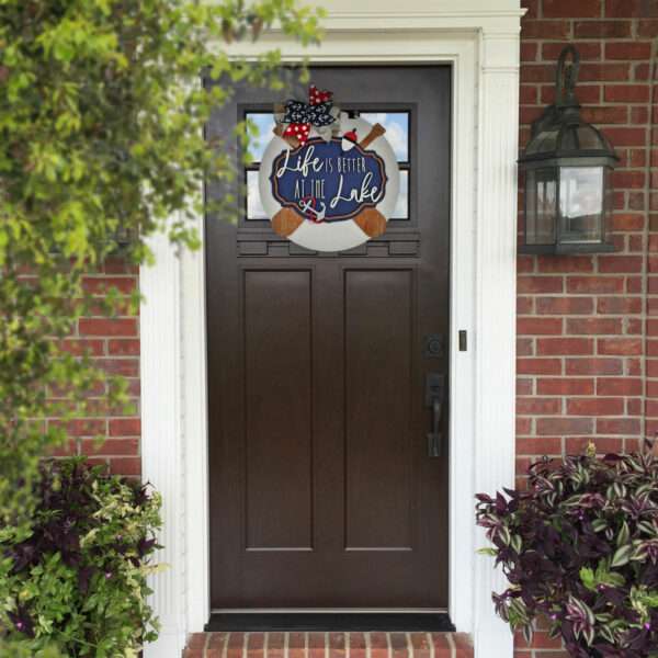 A front door with a custom decorative sign reading "Life is better at the lake," adorned with an anchor and life ring, hangs above. The door is surrounded by brick, and nearby plants add greenery. There's also a lantern-style light fixture affixed to the wall next to the door.