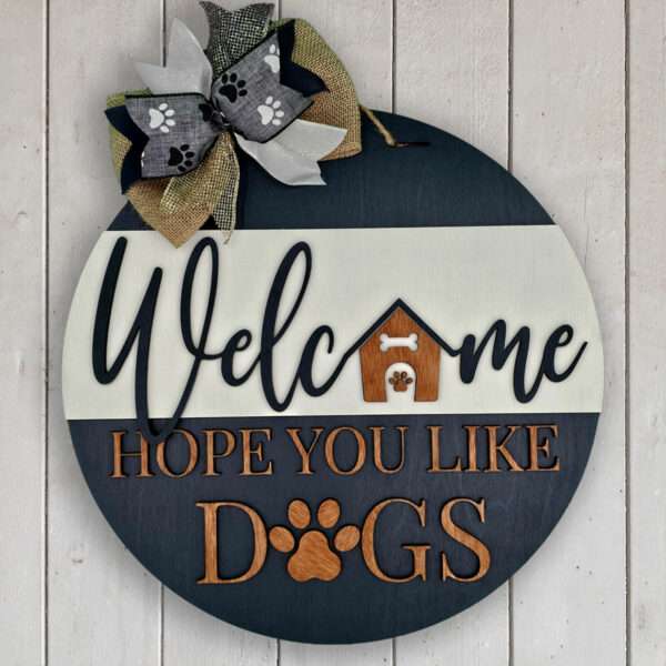 A round wooden welcome sign hangs against a white wall. The text, "Welcome, Hope You Like Dogs," is accented with paw prints and a dog house image. A patterned ribbon bow at the top adds charm, adorned with even more paw prints.