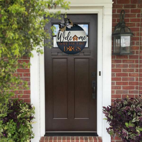 A dark brown front door with a glass panel at the top, set in a brick wall, surrounded by white trim. Above the door, a custom door sign reads, "Welcome, Hope You Like Dogs," with dog paw prints and a small house illustration. A garden lantern is on the right wall.